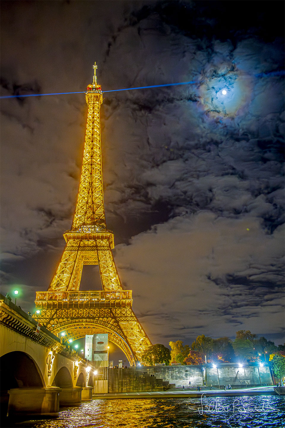 左图为法国巴黎著名的埃菲尔铁塔，在夜晚被金色的灯光照亮。顶部发出蓝色激光。云朵点缀着背景的天空。透过云层也可以看到月亮，但它被五颜六色的光环环绕着：月华。有关更多详细信息，请参阅说明。