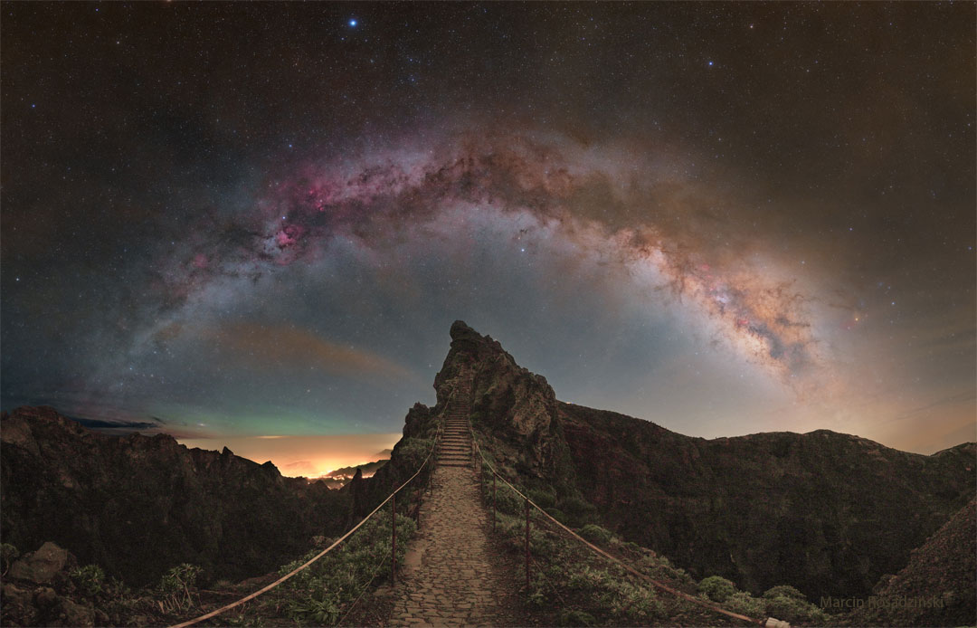 星光闪烁的天空显示出银河系中央带的拱形横跨图片顶部。前景是一片岩石景观，前方有一座小山，还有一条通往山上楼梯的小路。有关更多详细信息，请参阅说明。