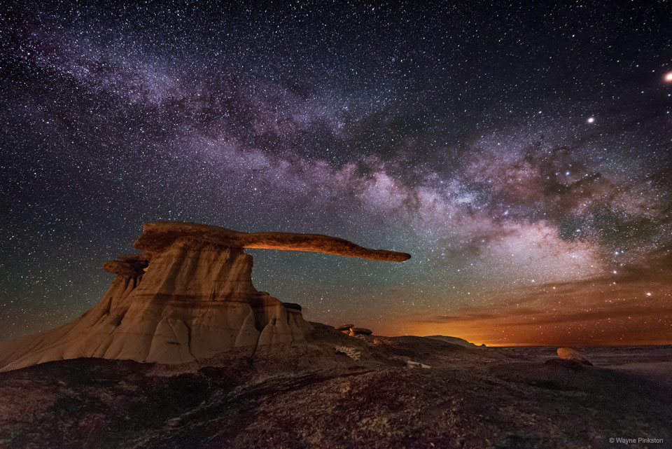 银河系盘面横跨繁星点点的夜空。右侧地平线上布满了彩云。图像中心出现了一个奇怪的岩石结构，其底部和延伸的臂状物似乎指向色彩斑斓的地平线。有关更多详细信息，请参阅说明。