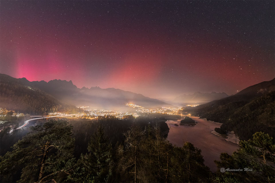 夜晚，城镇上方出现星空。天空左侧出现粉红色辉光，这是极光，而天空右侧出现更平滑、更暗的辉光，这是稳定极光红弧。有关更多详细信息，请参阅说明。